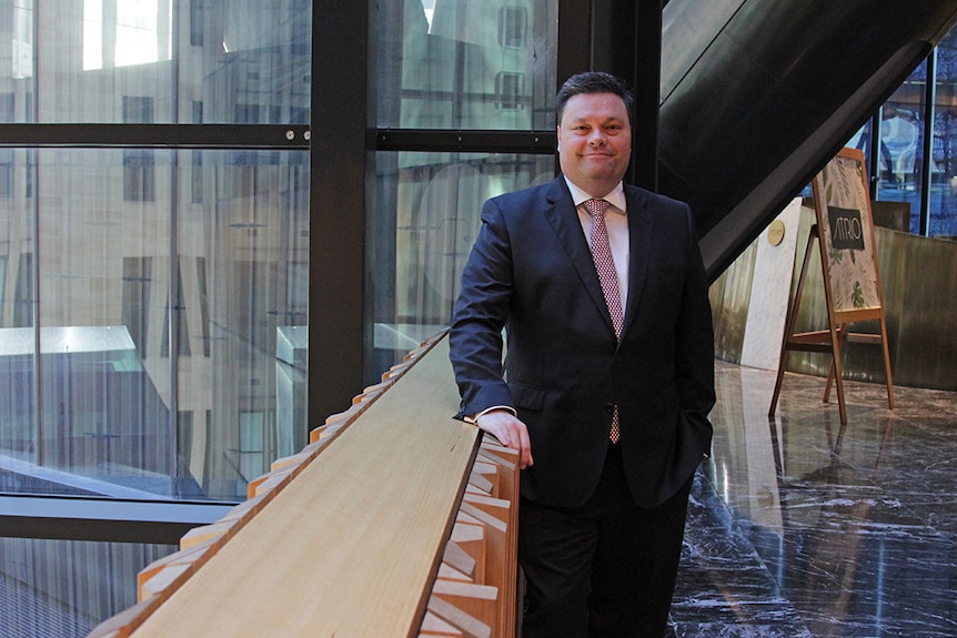 A man in a suit stands in an office building, smiling.