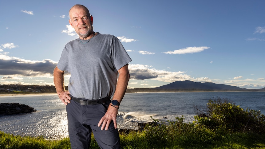 Middle-aged man standing on the edge of a beautiful oceanic hinterland, bald, grey stubble, grey tee, blue pants, slight smile.