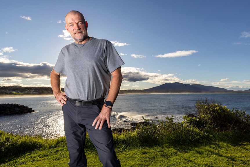 Middle-aged man standing on the edge of a beautiful oceanic hinterland, bald, grey stubble, grey tee, blue pants, slight smile.