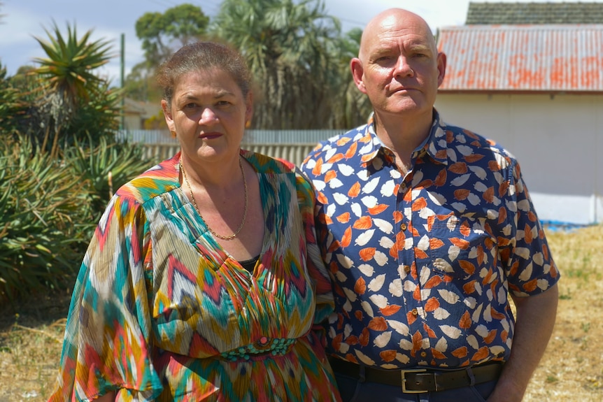 A woman and man standing in a grass field, looking at the camera with serious faces.