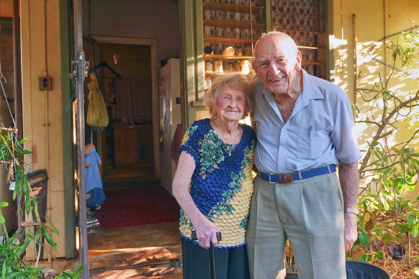 An elderly couple stand in front of their home.