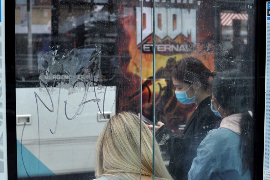 Two women wearing face masks stand at a bus stop. Behind them is a bus poster with the word 'Doom' in large letters.