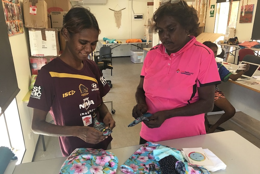 Two Domadgee women standing next to each other at a table while making Moon Sick car Bags.