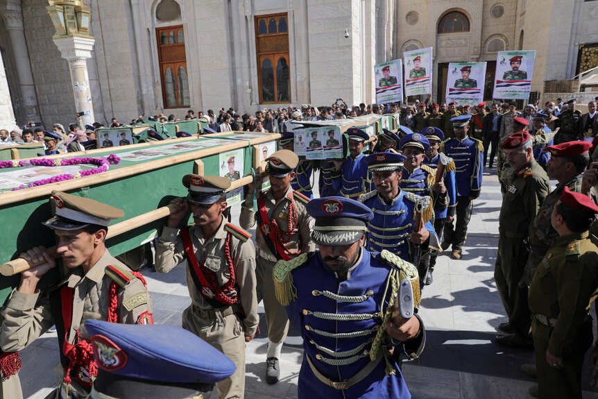 A wide shot of military guards carrying coffins in a line outside of a large building as a crowd watches on.