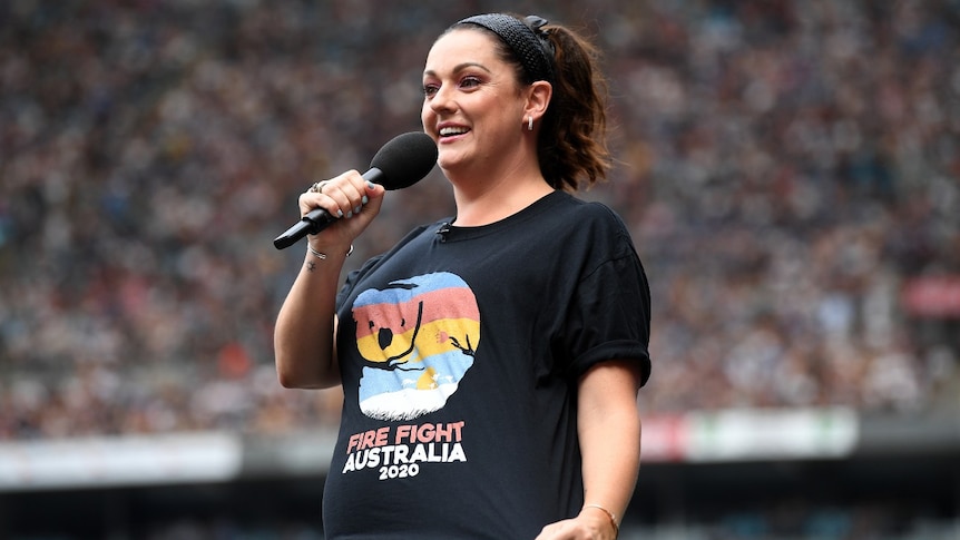 A woman holding a microphone shows a stadium crowd her Fire Fight Australia concert t-shirt.