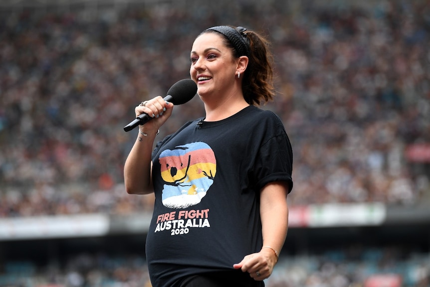 Une femme tenant un microphone montre à la foule du stade son t-shirt de concert Fire Fight Australia.
