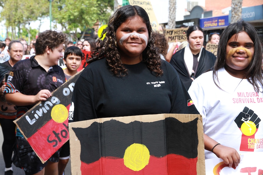 An Indigenous woman with white paint on her face holding a sign with an Aboriginal flag. 