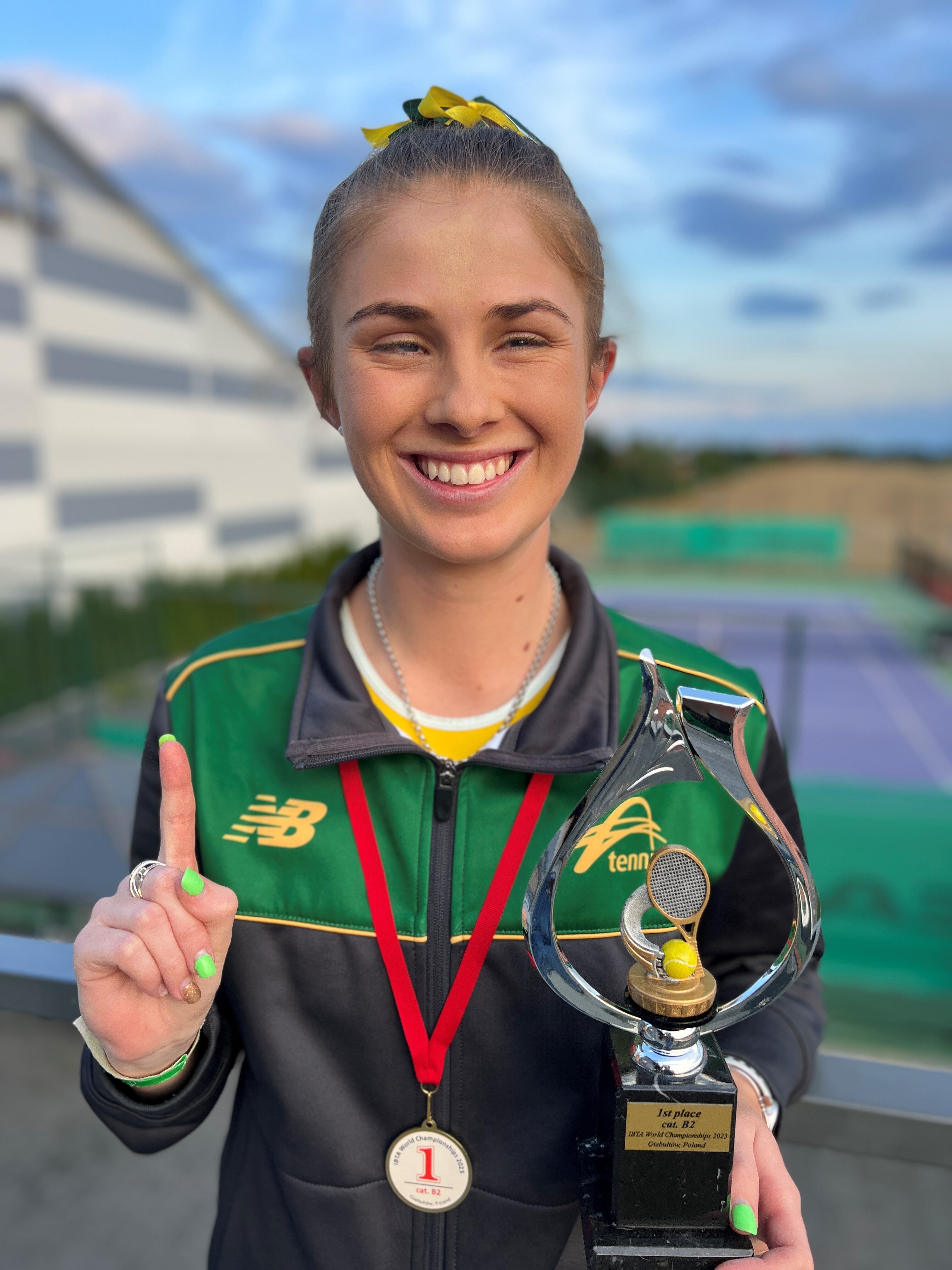 A young woman stands in a green and gold jacket holding a trophy and holding up one finger as a winner.