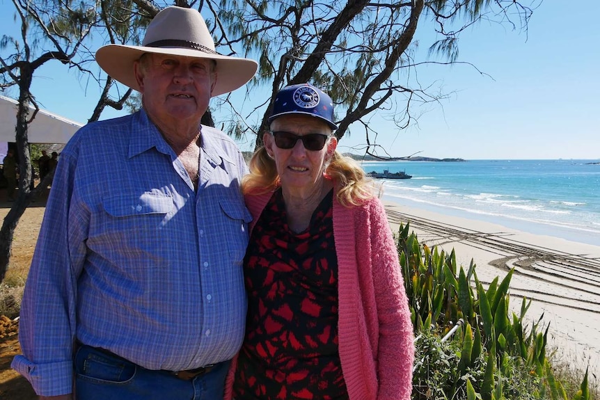 A man and woman stand facing the camera with blue ocean behind them and war ships coming into shore.