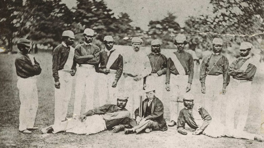 Twelve men in cricket uniforms pose for a photo in front of trees.