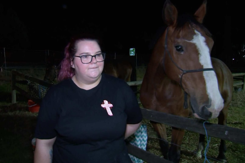 A woman wearing a black T-shirt with a cross on it with a horse at night
