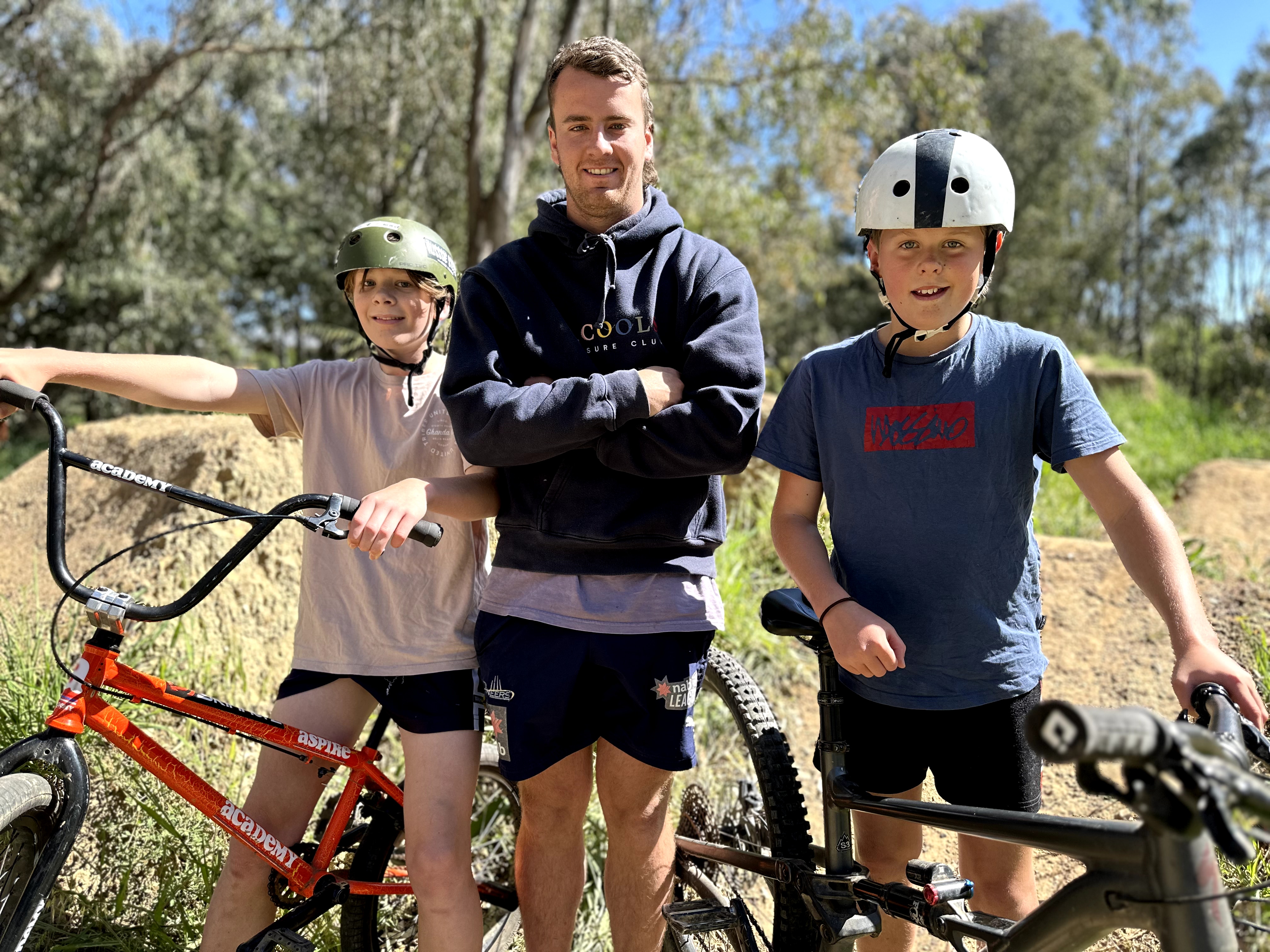 Two boys and a man stand in front of a dirt jump BMX track