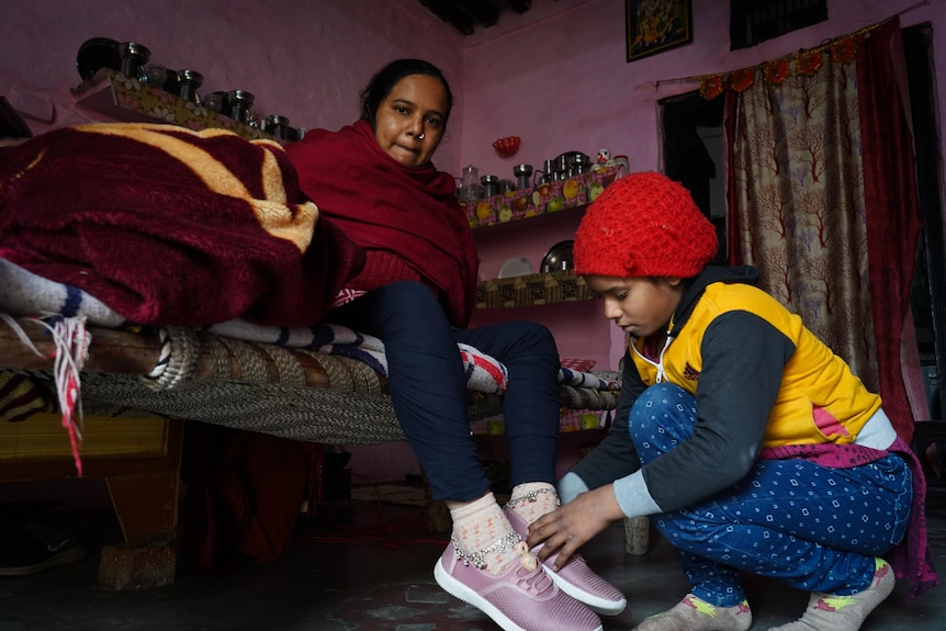 A woman sits on a bed while her child bends down to tie her shoelace