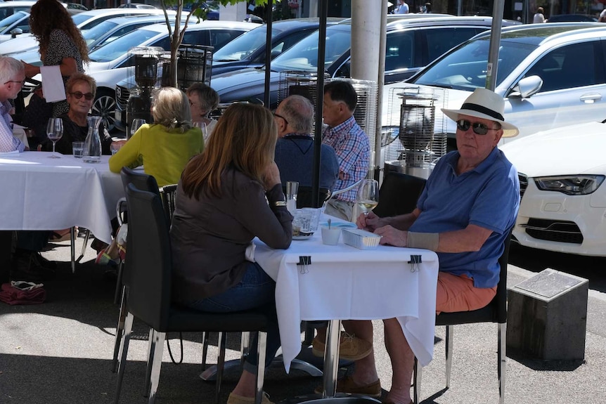 A number of groups of people eat outdoors at restaurants under large umbrellas.