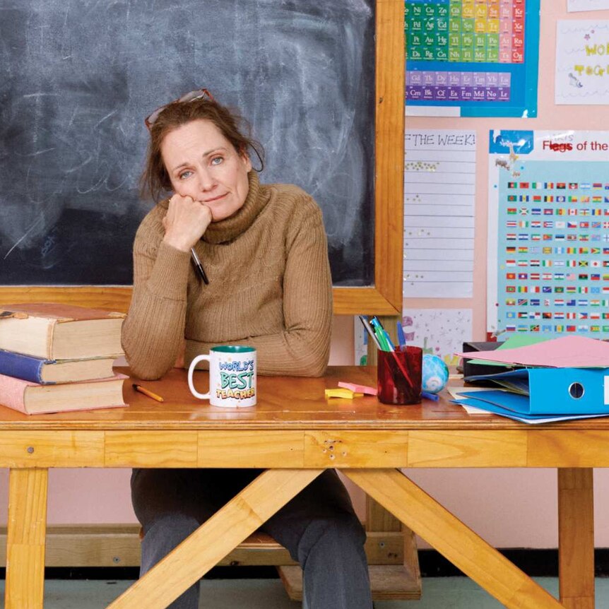 Catherine McClements, looking exhausted, sits at a desk in front of a blackboard.