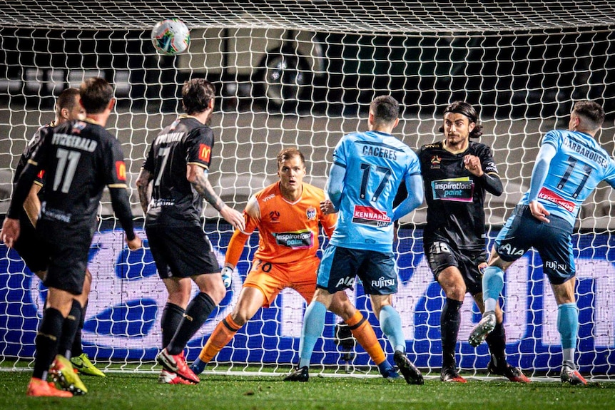 A goalkeeper stares wide-eyed at a ball in front of him. A Sydney FC player looks to have just completed a header