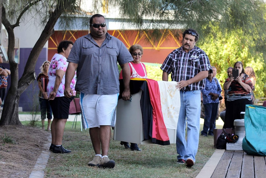 Two Aboriginal men carry a box draped in the Aboriginal flag.