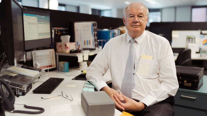 Brian Childs sits at his desk and computer in an office looking to the camera wearing a white business shirt and striped tie.