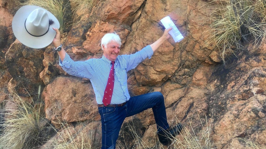 Bob Katter holds an Akubra in one hand and a letter in the other, arms up, smiling, with one leg leaning on a big red rock