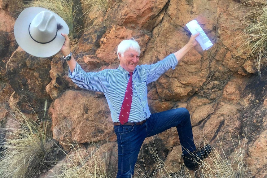 Bob Katter holds an Akubra in one hand and a letter in the other, arms up, smiling, with one leg leaning on a big red rock.