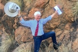 Bob Katter holds an Akubra in one hand and a letter in the other, arms up, smiling, with one leg leaning on a big red rock