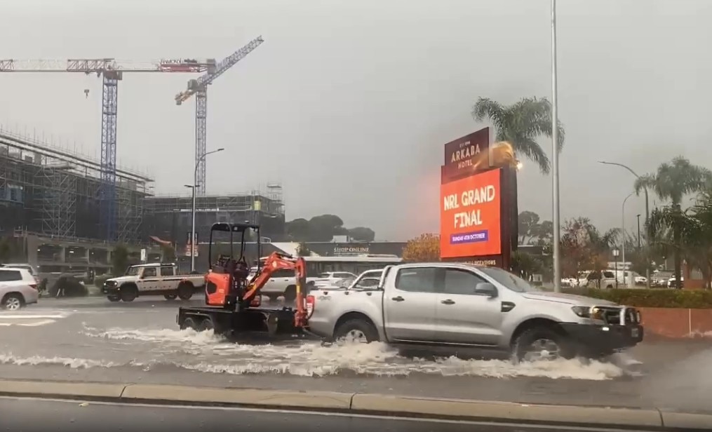 A ute trailer goes through a large amount of water on a road with a sign for NRL Grand Final behind