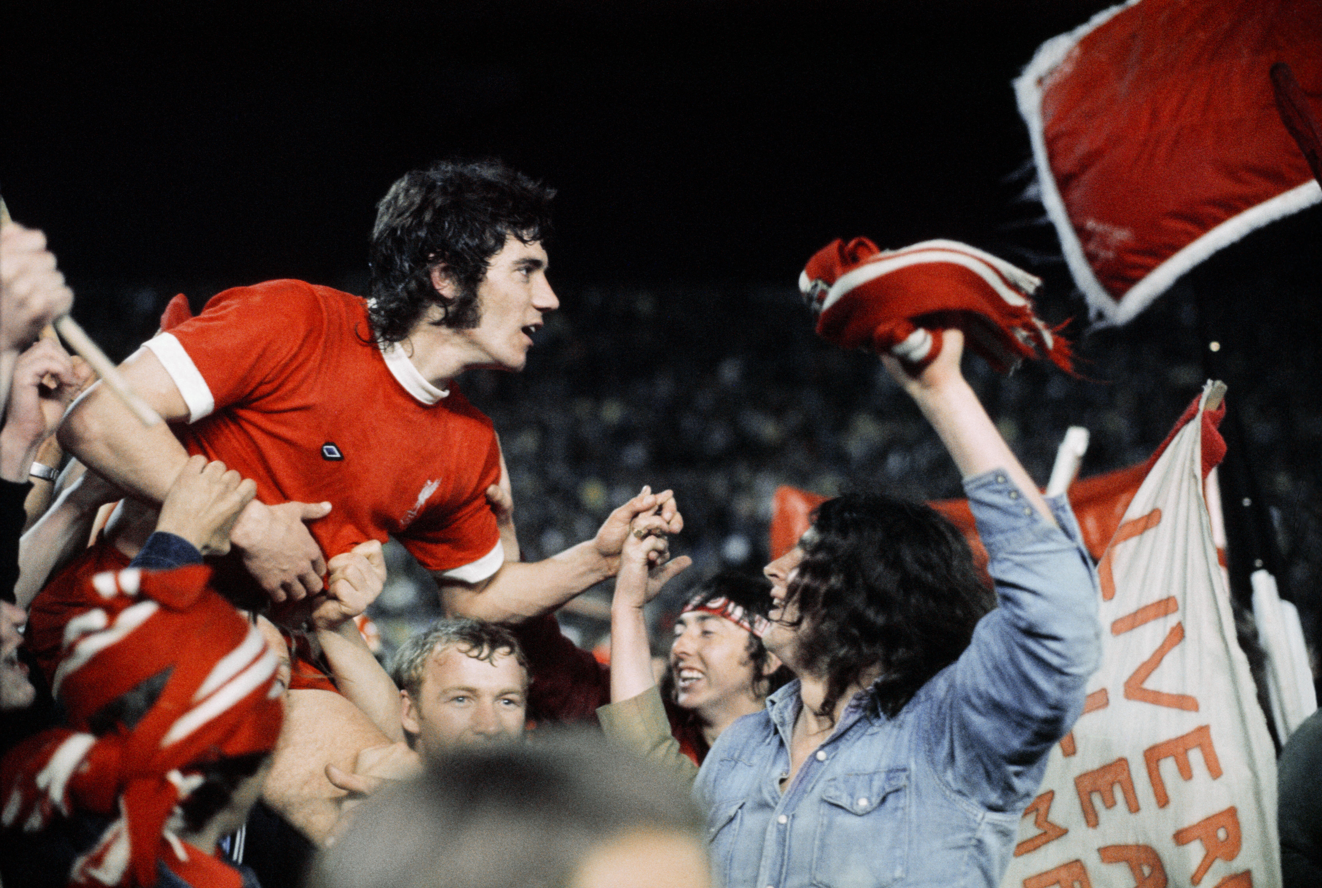 Celebrating fans carry a dark-haired player in a red shirt and with a 70s hairstyle and sideburns.
