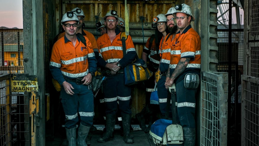 six miners in an elevator, wearing orange overalls, in a lift