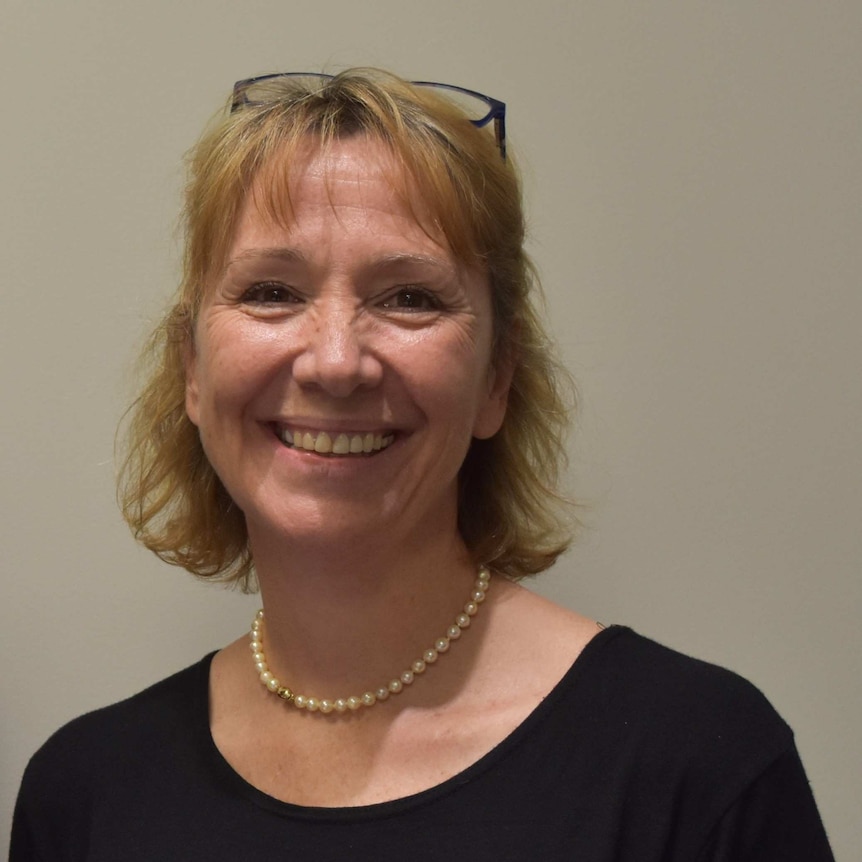 A woman with mid-length blonde hair and black reading glasses on her head smiling