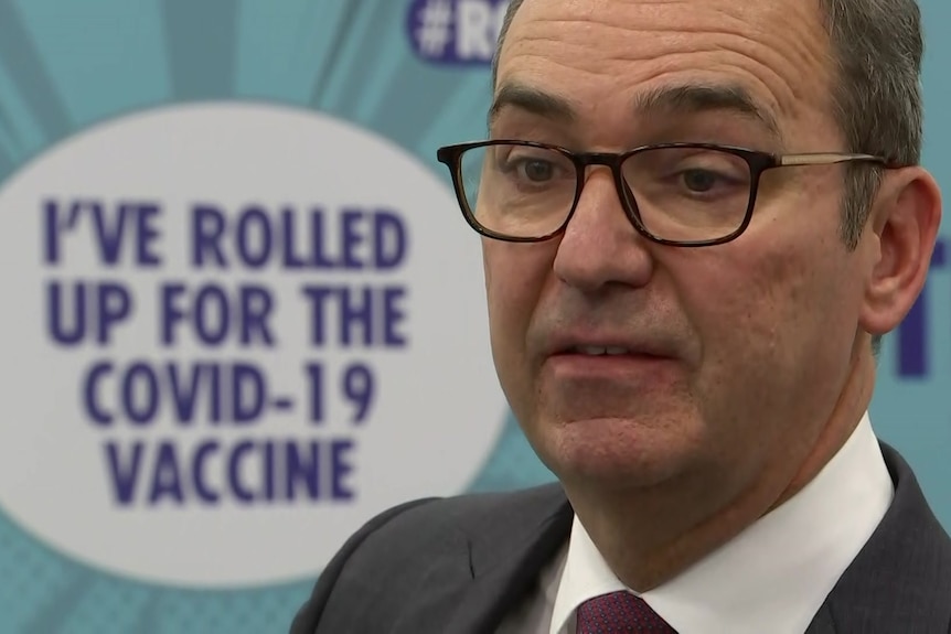 A man with glasses and dark hair stands in front of a sign saying I've rolled up for the COVID–19 vaccine