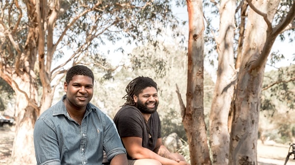 Two men seated, smiling at the camera, with gum trees in background.