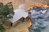 A still image from aerial footage shows flames burning towards a brick shed with a tin roof on a rural property.