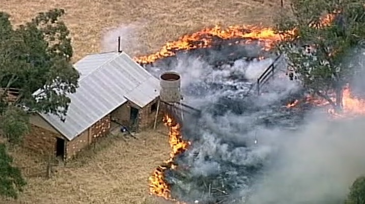 A still image from aerial footage shows flames burning towards a brick shed with a tin roof on a rural property.