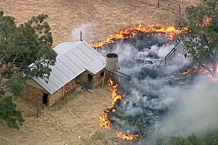 A still image from aerial footage shows flames burning towards a brick shed with a tin roof on a rural property.