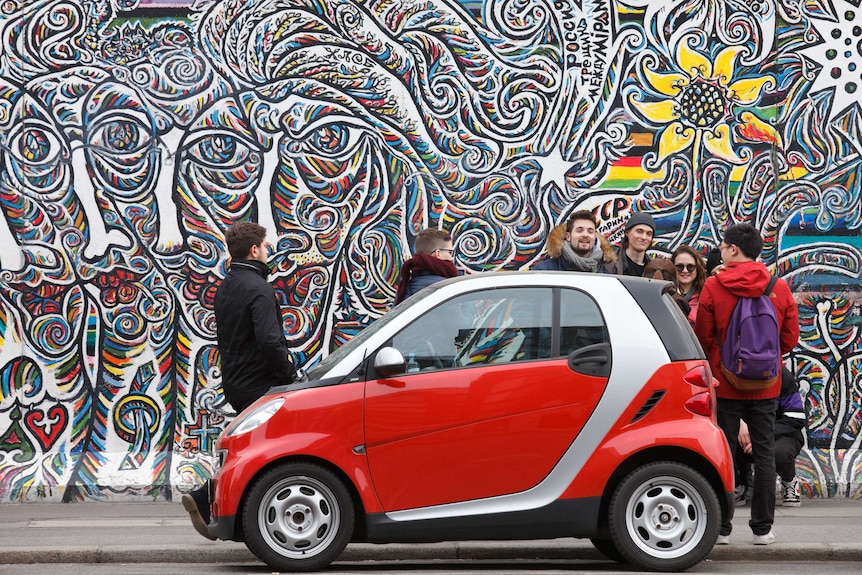 A small red car and a group of people in front of a mural on the Berlin Wall.