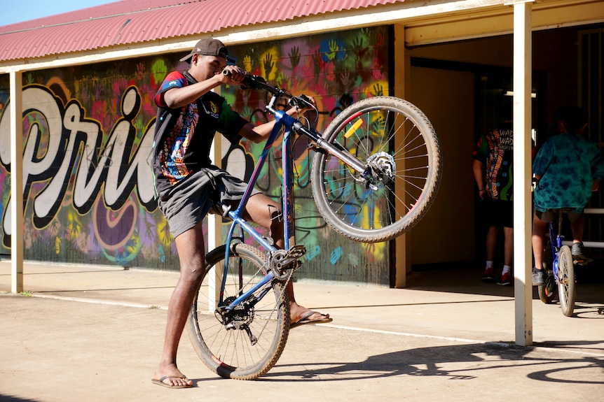 a young man on a bike, front tyre in the air, hands on handle bars, one foot on the ground, colourful wall behind.