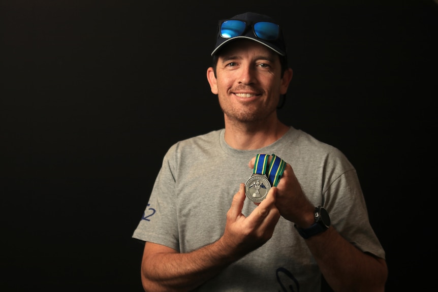 A young man wears a grey t-shirt and blue hat and holds a silver medal, smiling.