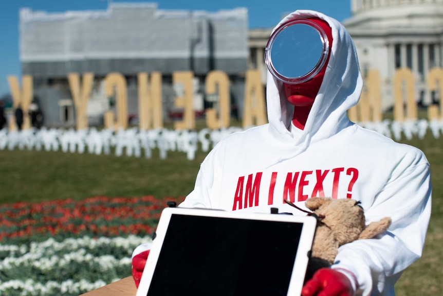 A dummy in a white sweatshirt and a mirror for a face sits at a school desk in a circle of roses on the US capitol lawn.