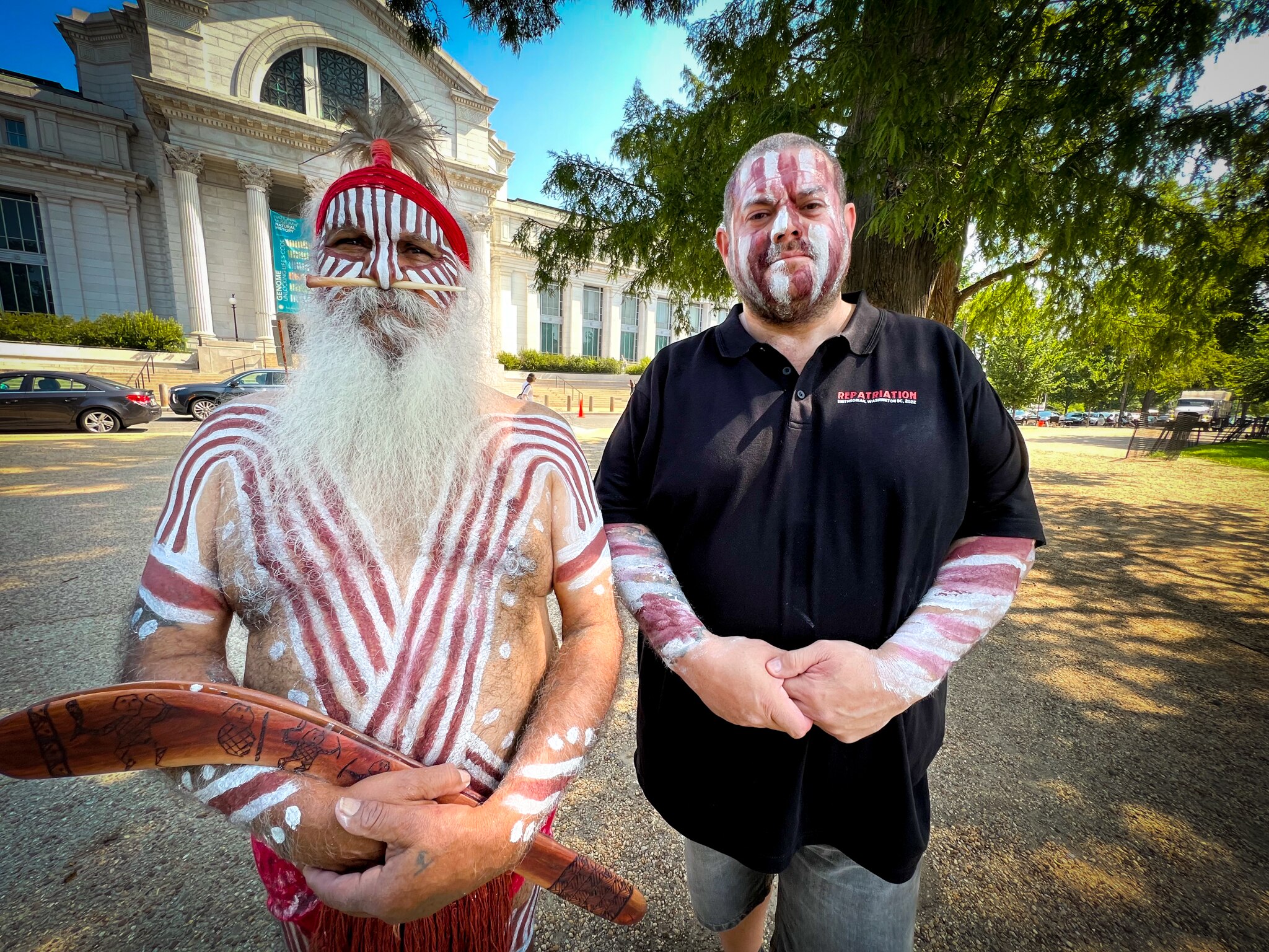 Two indigenous men, one wearing traditional dress and holding a boomerang, the other in black polo, stand side by side