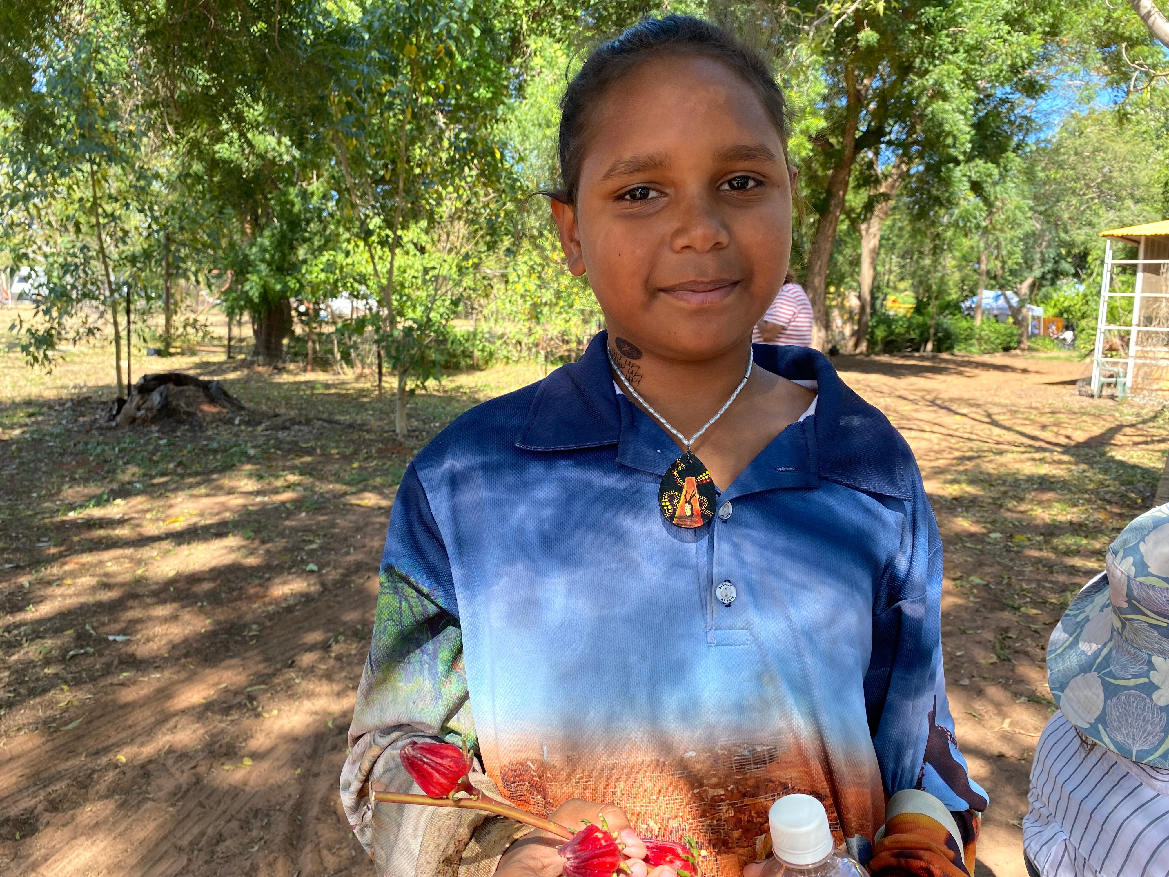 A young girl in a blue and orange long sleeve shirt smiles as she holds a branch of rosella