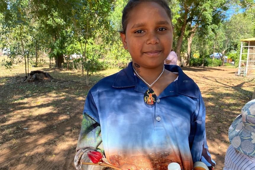 A young girl in a blue and orange long sleeve shirt smiles as she holds a branch of rosella