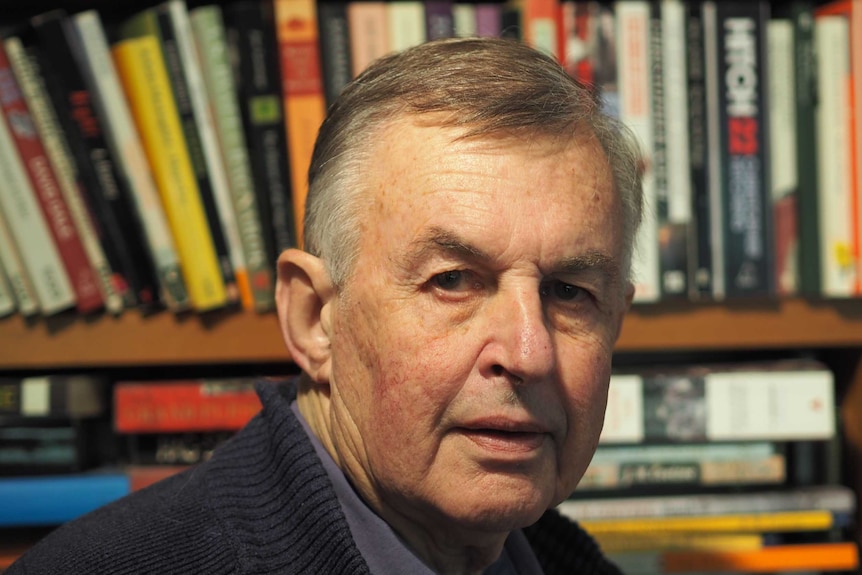 A grey haired man stares at the camera with many books in a shelf in the background.