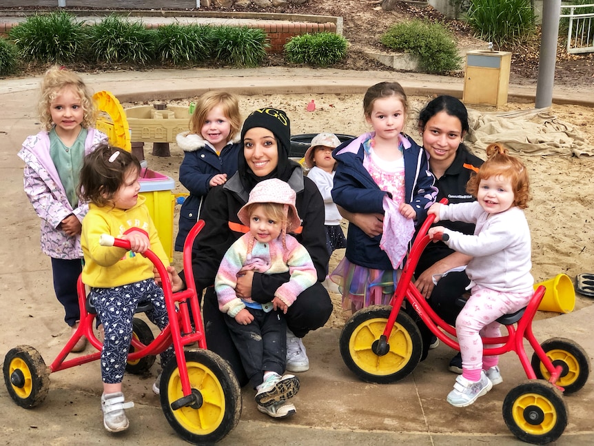 Two women with a number of children. Two of the children are riding tricycles.