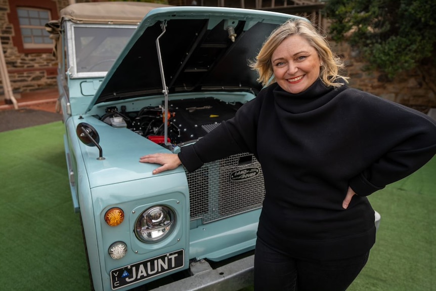 Lady smiling with blonde hair standing infront of blue vehicle 