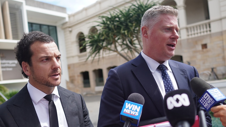 Man wears dark suit and tie and stares seriously towards reporters standing out of frame