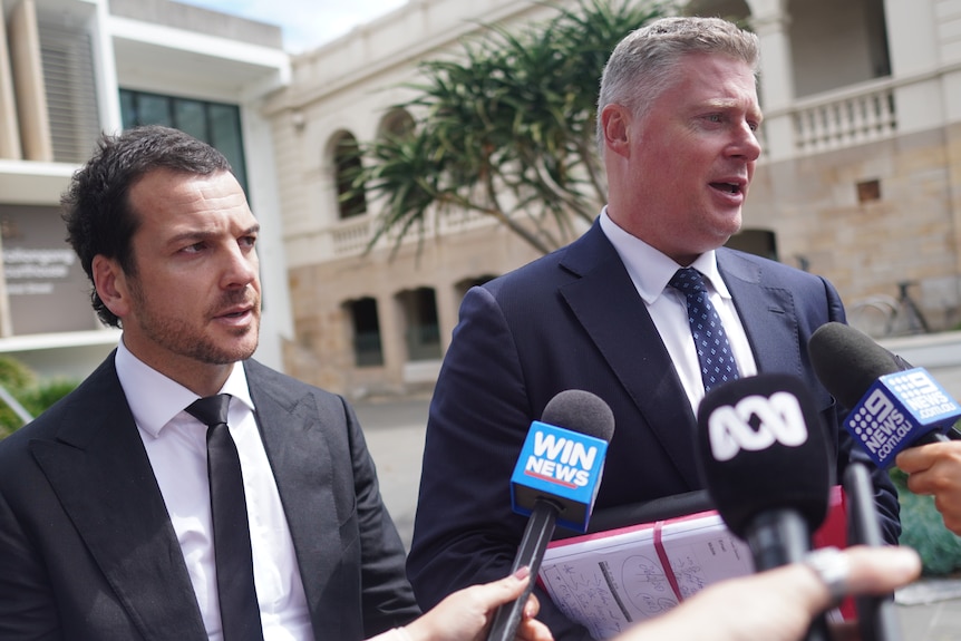 Man wears dark suit and tie and stares seriously towards reporters standing out of frame