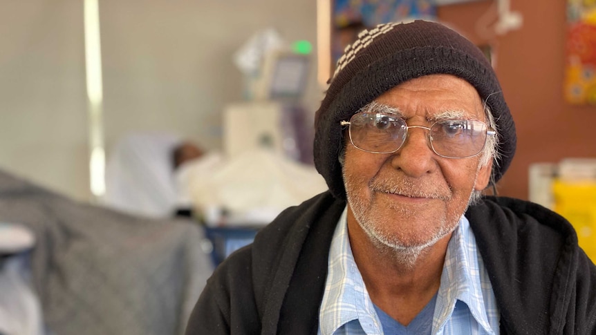 A man in glasses looks down the camera with a slight smile wearing a beanie and blue shirt