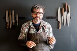 Artisan knife maker Todd Neale in his workshop standing in front of eight knives mounted on the wall.