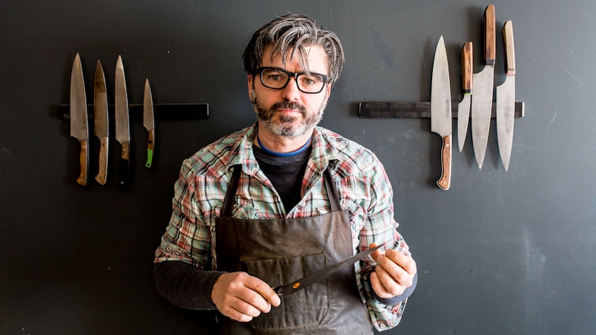 Artisan knife maker Todd Neale in his workshop standing in front of eight knives mounted on the wall.