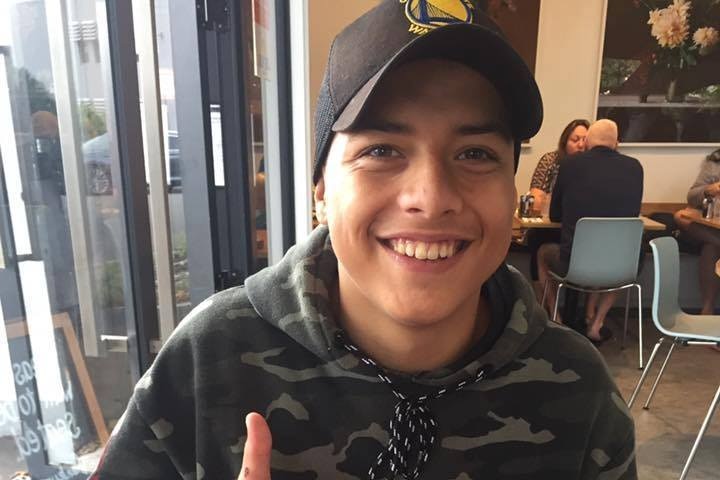 A young man in a camo jumper smiles happily at the camera in a cafe.